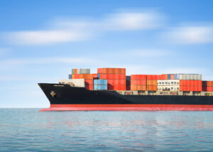 A large cargo ship loaded with stacked shipping containers sails on calm blue water under a clear sky, showcasing how to minimize damage while shipping cargo through secure stacking and smooth sailing conditions.