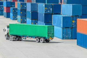 A white semi-truck carries a long green shipping container past stacks of colorful cargo—blue, red, and orange—at a shipping yard, showcasing how to minimize damage while shipping cargo through organized container management.