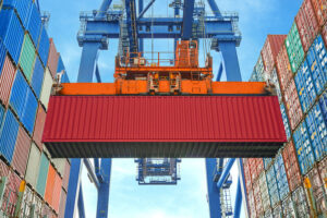 A large red shipping container is being lifted by an orange crane at a busy port, surrounded by stacks of colorful containers under a blue sky—ideal conditions to avoid condensation in a shipping container.