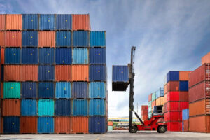 A large red forklift moves a shipping container in a yard filled with tall stacks of colorful cargo containers under a blue sky, highlighting the importance of knowing how to keep shipping containers cool in sunny conditions.