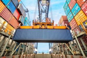 A large crane lifts a shipping container over rows of stacked colorful containers at a busy industrial port, where the benefits of using shipping container desiccants help protect cargo under the clear blue sky.