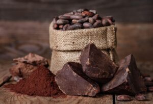 Large chunks of dark chocolate, cocoa powder, and a burlap sack filled with cocoa beans—freshly Cocoa Transported—are arranged on a rustic wooden surface, with more cocoa beans scattered around.