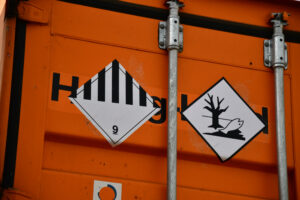 Close-up of an orange metal container with two hazard signs, including one for environmental hazards—a crucial reminder about how to ship dangerous chemicals safely. Vertical metal bars run over the black-and-white-striped and wildlife warning signs.