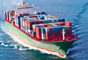 A large cargo ship carrying numerous colorful shipping containers sails through the blue ocean, creating white waves as it moves forward. Ever wondered how hot do shipping containers get under the sun during such voyages?.