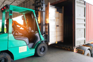 A person operates a green forklift, loading a stack of boxes wrapped in plastic into a shipping container on a truck inside a warehouse, highlighting the benefits of using shipping container desiccants for protecting goods during transit.