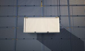 A white shipping container is being lifted by a crane in front of a stack of blue containers, casting a diagonal shadow—an example of how to keep shipping container cool by using shade and strategic placement.