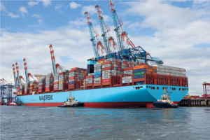 A large Maersk container ship loaded with colorful shipping containers is docked at a port, surrounded by cranes and two tugboats on the water, under a partly cloudy sky—a scene reflecting the shipping industrys role in climate change.