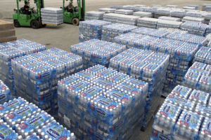 Stacks of bottled water cases are arranged in rows outdoors, with two green forklifts in the background transporting more cases. The pallets are shrink-wrapped and labeled Dannon—ideal conditions to consider: What is a thermal pallet cover?.
