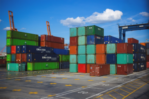 Stacks of colorful shipping containers are arranged in an orderly fashion at a shipping port, with large cranes overhead and a blue sky dotted with clouds. The ground is marked with numbered parking spots, highlighting where items like thermal pallet covers might be stored.