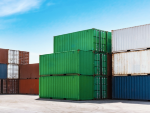 A stack of multicolored shipping containers, including green, brown, white, and blue, sits on a concrete lot under a clear blue sky—showcasing global hygiene standards to help prevent mold issues.