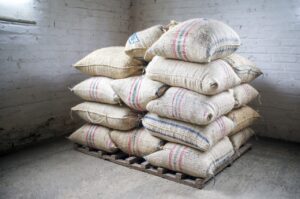 A stack of burlap sacks with red and blue stripes, ideal for Protecting Your Exports, rests on a wooden pallet in the corner of a white brick room with concrete floors. Light comes in through a small window on the left.
