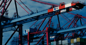 Large red and blue cranes stand over stacked shipping containers at a busy port, with a partly cloudy sky in the background. Signs on the cranes read EUROGATE Container Terminal.