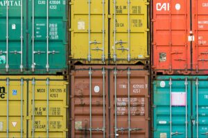 A close-up view of stacked shipping containers in various colors, featuring visible metal bars, hinges, white identification markings, and some fitted with Thermal Container Liners to help protect temperature-sensitive cargo.
