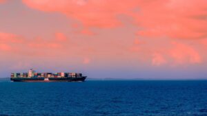 A large cargo ship loaded with colorful thermal container liners sails on the deep blue ocean under a sky filled with dramatic pink clouds at sunset.