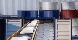 Conveyor belt transporting white sacks into an open shipping container, with additional stacked containers in the background.