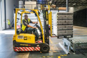 A warehouse worker in a yellow safety vest operates a yellow Hyster forklift, moving boxed goods onto a conveyor system inside a large industrial warehouse, where protocols like how to prevent mold in shipping containers are carefully followed.