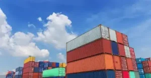 Stacks of colorful shipping containers are piled high under a bright blue sky with scattered white clouds, hinting at an active shipping yard where recent container shipping delays may be affecting operations.