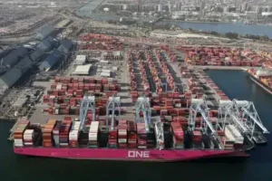 Aerial view of a busy shipping port with large cranes unloading containers from a pink cargo ship labeled ONE. Numerous shipping containers, hinting at container shipping delays, are stacked across the dock, with city and industrial buildings in the background.