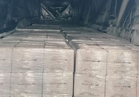 Stacks of large, shrink-wrapped boxes are lined up inside a dark trailer or storage container, filling the space from floor to ceiling.